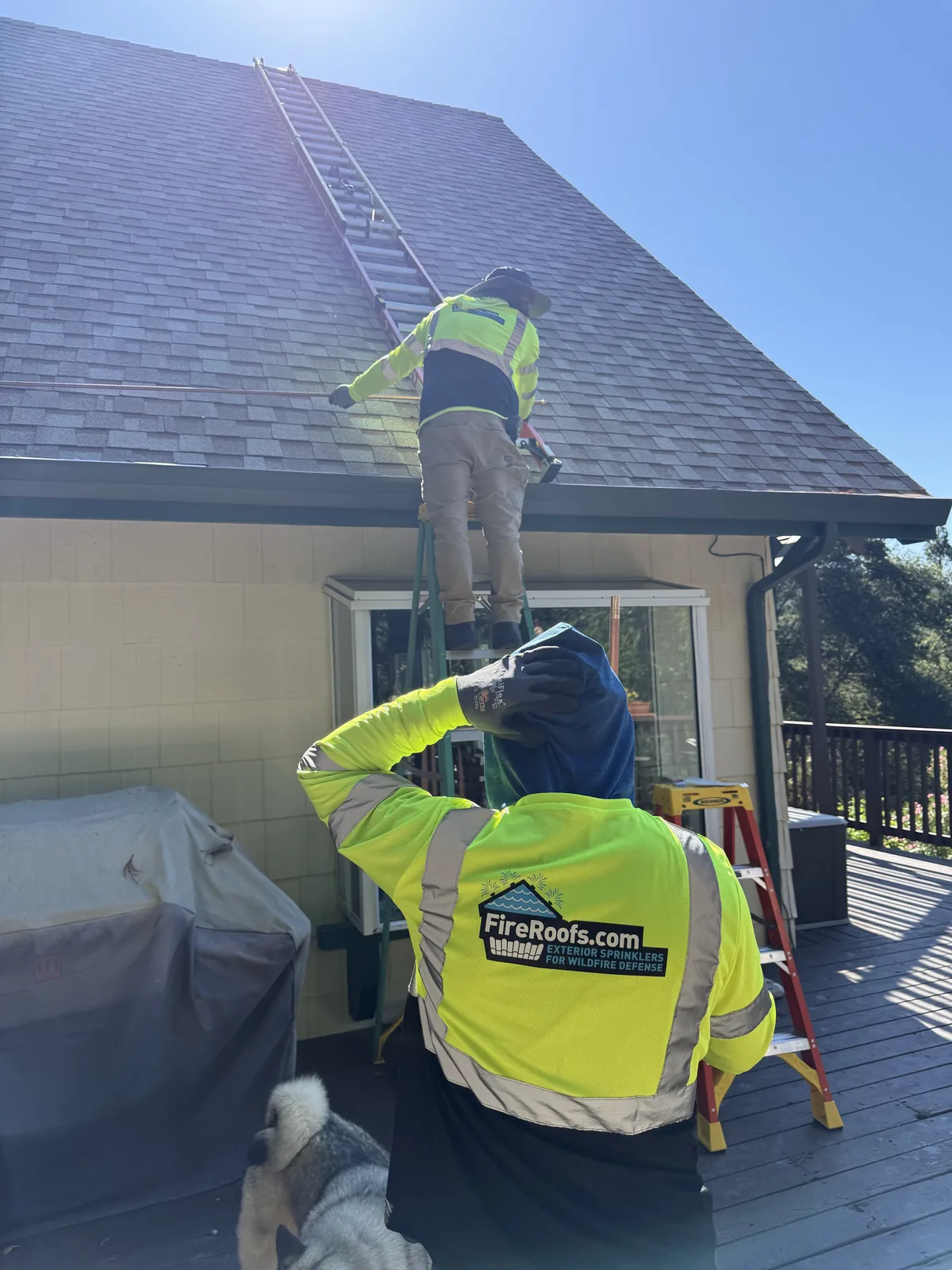 FireRoofs crew in branded high-visibility vests installing exterior wildfire sprinklers from a ladder on a Bay Area home in wooded Santa Cruz Mountains terrain