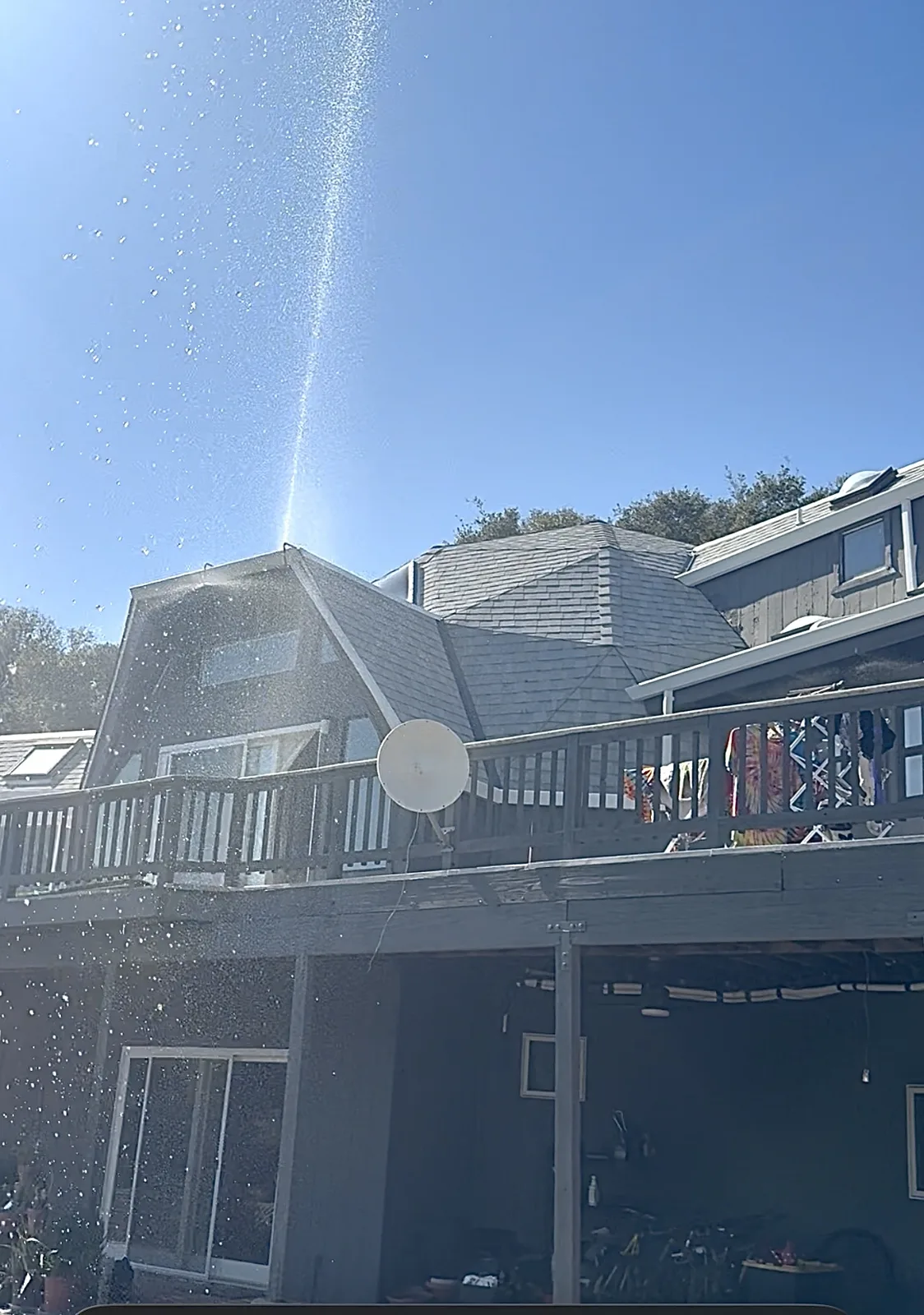 A FireRoofs roof sprinkler throwing a tall column of water above a Bay Area hillside home with a wraparound deck